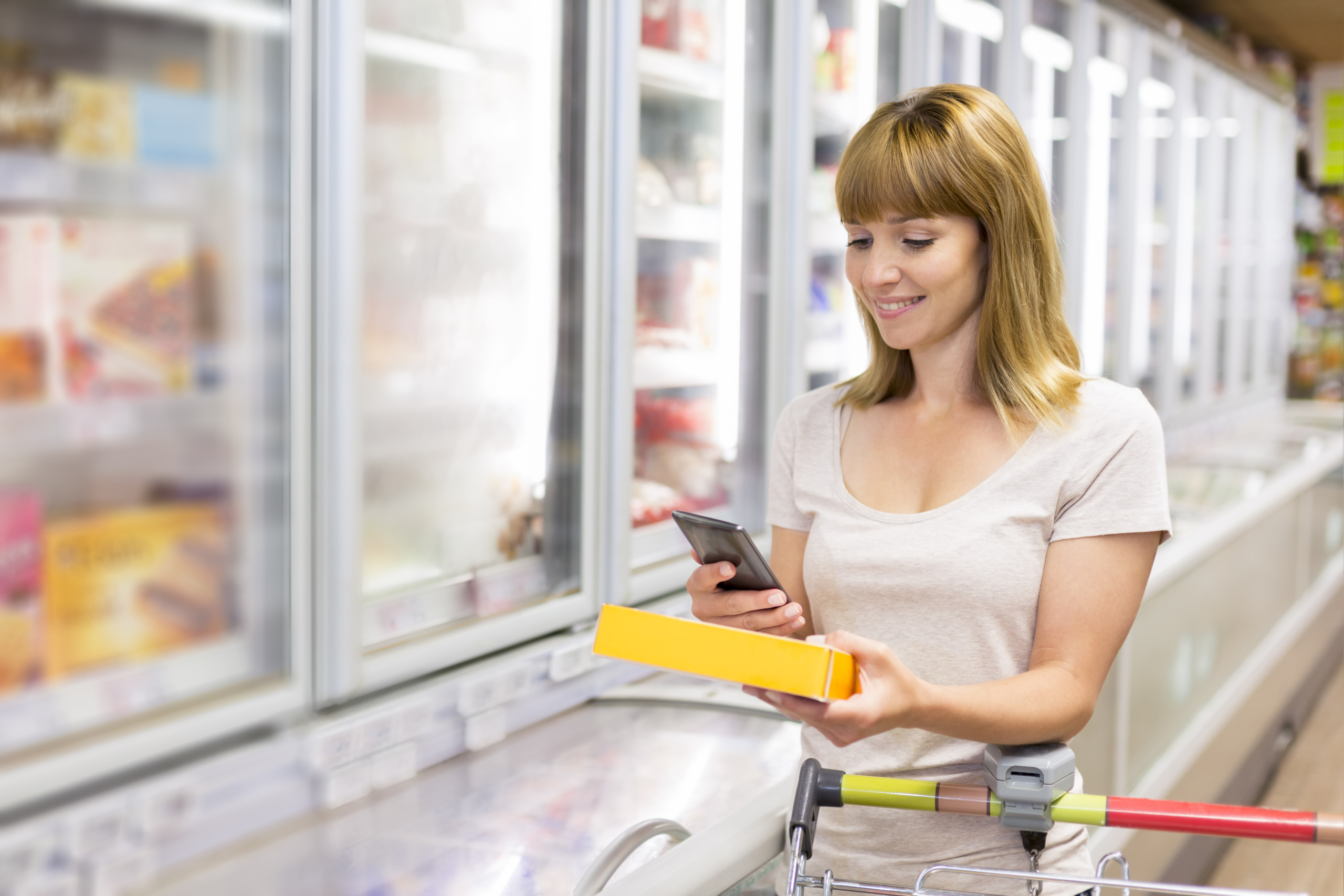 Woman in a supermarket aisle behind a trolley holding a product and lookign at her phone